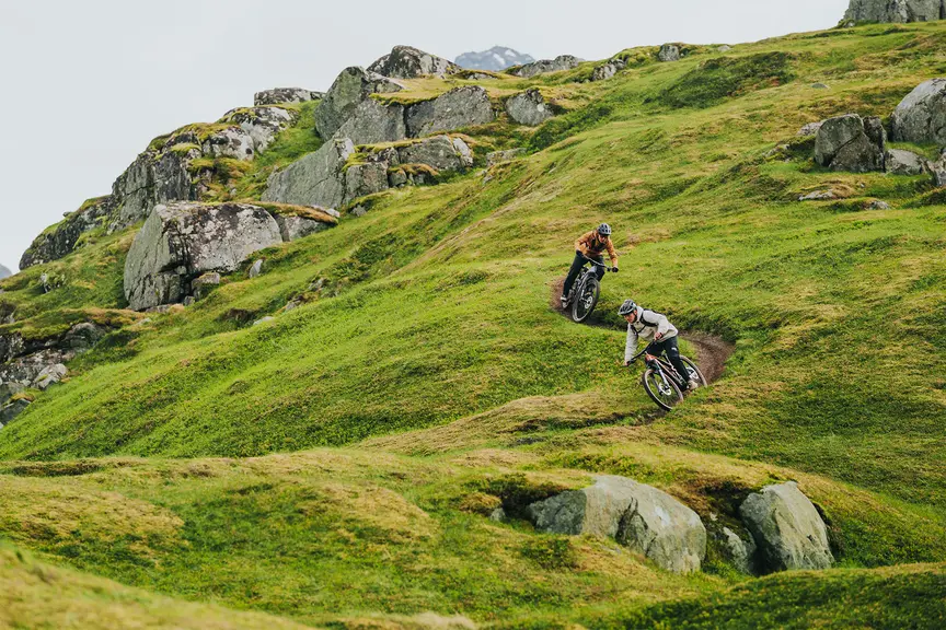 Twee mensen rijden op e-mountainbikes over een smal pad door een groen, heuvelachtig landschap. De omgeving is bergachtig met glooiende heuvels en grote rotsen. Beide mensen dragen helmen en buitenkleding.