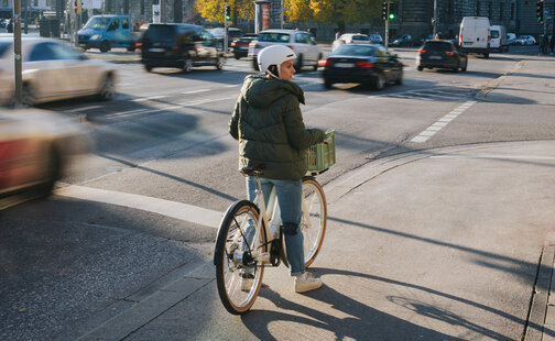 Une femme se tient à un carrefour avec son vélo à assistance électrique et regarde vers la droite. À l'arrière-plan, on peut voir des voitures et des feux de signalisation.