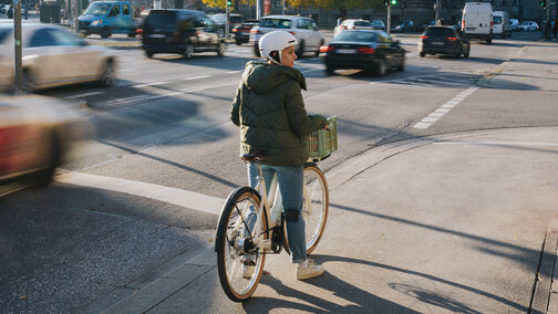 A woman is standing with her eBike at an intersection, looking to the right. In the background, there are some cars and traffic lights visible.
