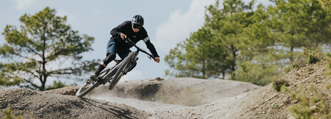 A man is riding quickly down an outdoor trail, with the front wheel of his eBike in the air. Trees are visible in the background.