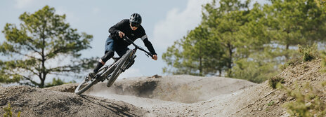 A man is riding quickly down an outdoor trail, with the front wheel of his eBike in the air. Trees are visible in the background.