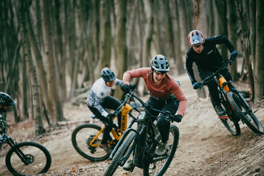 Vier Personen fahren auf eMountainbikes hintereinander einen erdigen Uphillpfad hinauf. Sie haben freudig ihre Münder aufgerissen. Im Hintergrund sieht man kalte Baumstämme und herabgefallene braune Blätter.