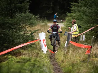 An eBike rider is seated on a mountain bike, with a person in a yellow safety vest standing next to them, holding a leaf in their hand. In front of them, there is a narrow trail delimited by red barrier tape, with trees visible in the background.