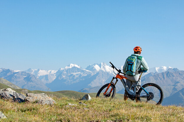 An eBike rider stands leaning against his eBike on a hill and looks into the distance at mountains.