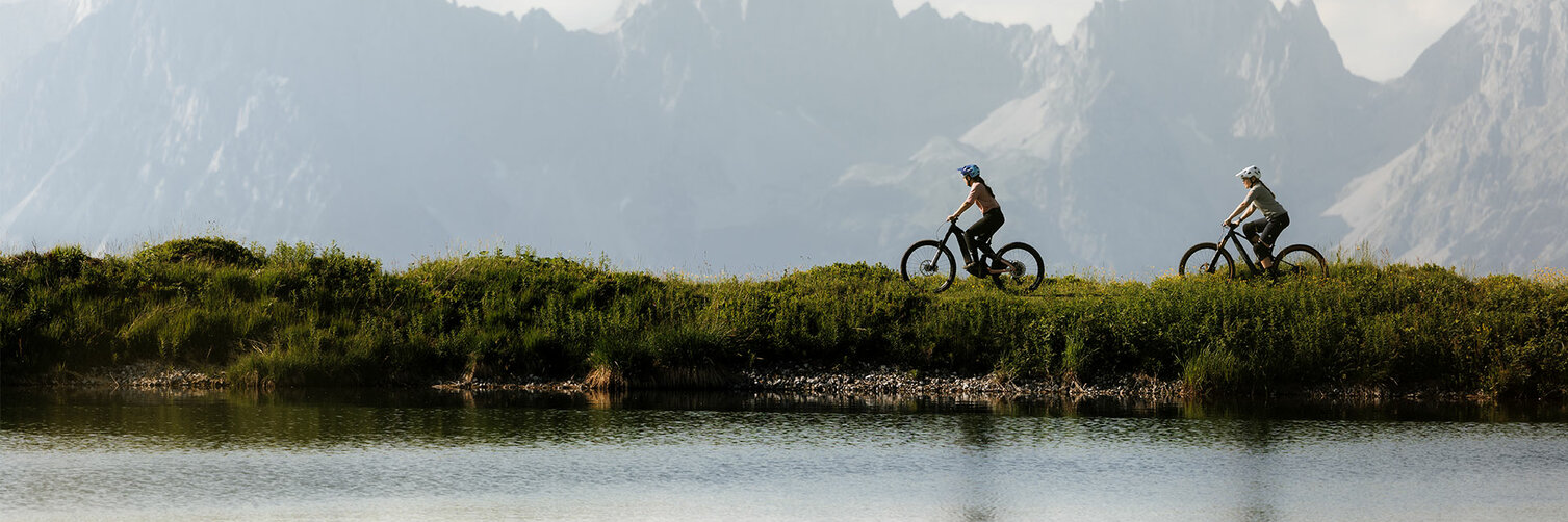 Blick auf einen See. Im Hintergrund fahren zwei Frauen auf ihren eBikes.