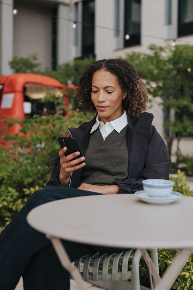 En kvinna sitter på ett utomhuscafé i staden och tittar på sin smartphone. Scenen fångar ett ögonblick av lugn och uppkoppling i det moderna vardagslivet.