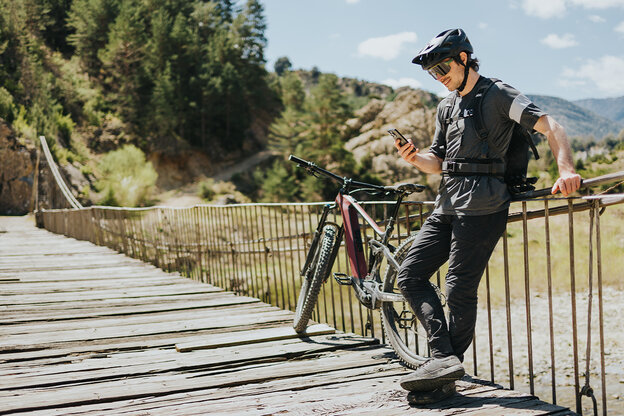 A eBike rider stands leaning against a railing on a bridge over a river and looks at his phone, next to him stands his eBike.