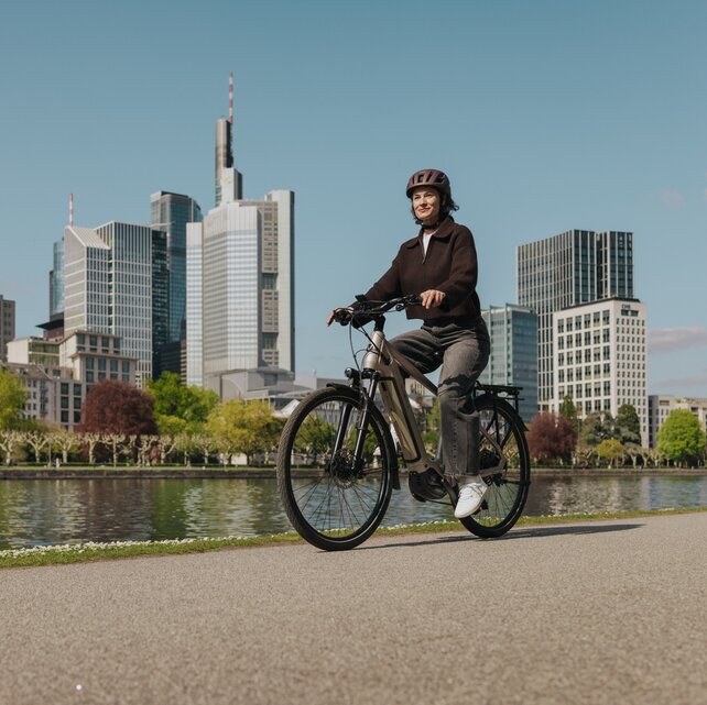 Eine Frau genießt eine Fahrt mit ihrem E-Bike auf einem Radweg am Flussufer, mit der Skyline von Frankfurt im Hintergrund.
