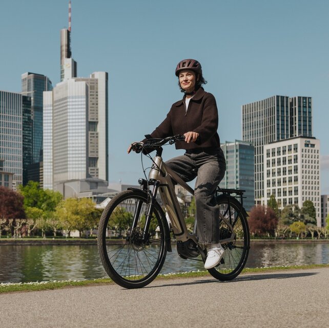Eine Frau genießt eine Fahrt mit ihrem E-Bike auf einem Radweg am Flussufer, mit der Skyline von Frankfurt im Hintergrund.