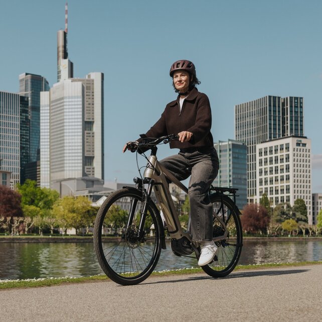 Eine Frau genießt eine Fahrt mit ihrem E-Bike auf einem Radweg am Flussufer, mit der Skyline von Frankfurt im Hintergrund.