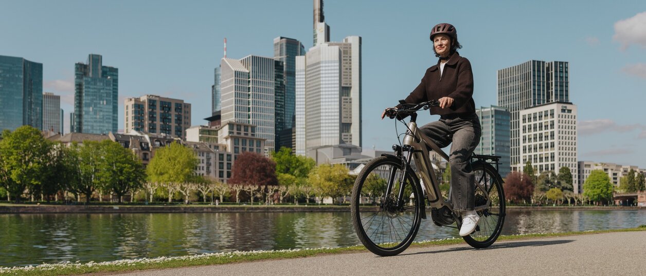 Une femme profite d'une balade en vélo électrique sur une piste cyclable au bord de la rivière, avec la silhouette de Francfort en arrière-plan.