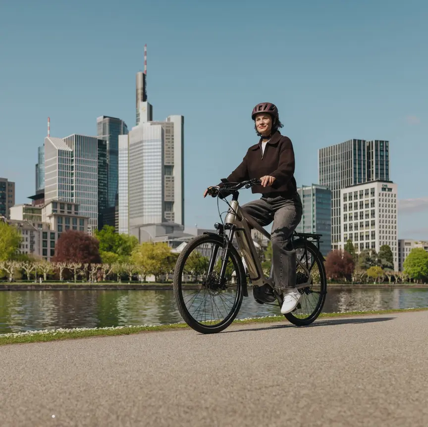 Eine Frau genießt eine Fahrt mit ihrem E-Bike auf einem Radweg am Flussufer, mit der Skyline von Frankfurt im Hintergrund.