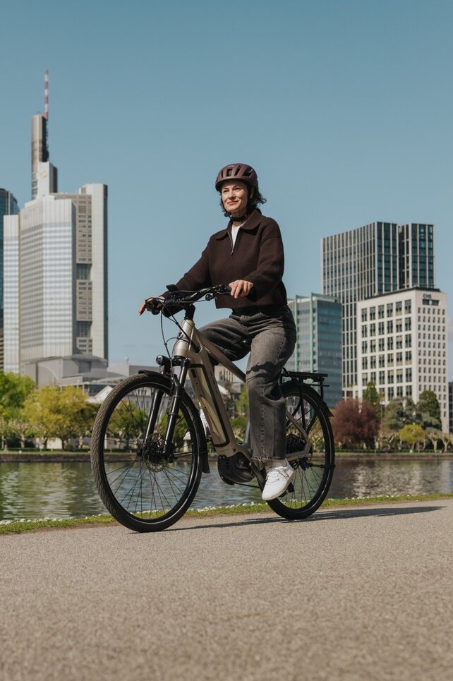 A woman enjoys a ride on her eBike along a riverside path, with the Frankfurt skyline in the background.