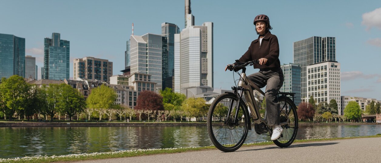 Eine Frau genießt eine Fahrt mit ihrem E-Bike auf einem Radweg am Flussufer, mit der Skyline von Frankfurt im Hintergrund.
