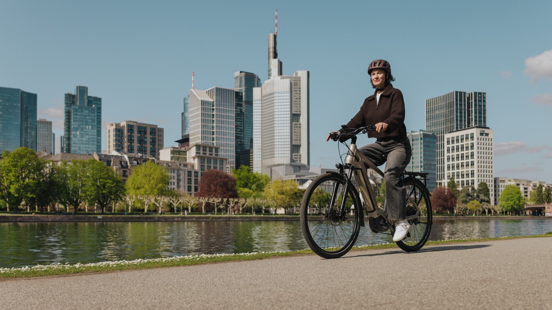 Eine Frau genießt eine Fahrt mit ihrem E-Bike auf einem Radweg am Flussufer, mit der Skyline von Frankfurt im Hintergrund.