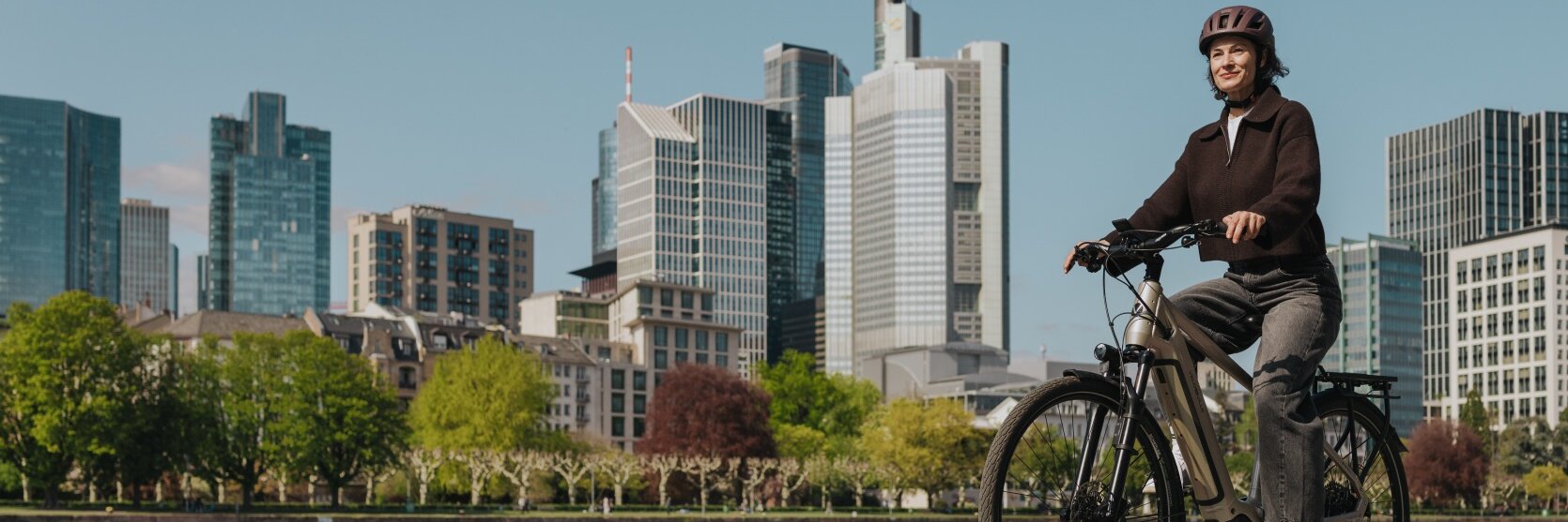 En kvinne nyter en tur på eBike-en sin på en sykkelsti langs elven, med Frankfurts silhuett i bakgrunnen.