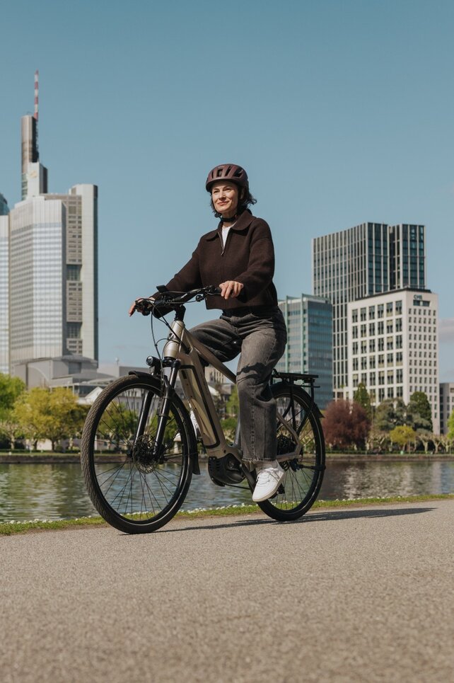 Eine Frau genießt eine Fahrt mit ihrem E-Bike auf einem Radweg am Flussufer, mit der Skyline von Frankfurt im Hintergrund.