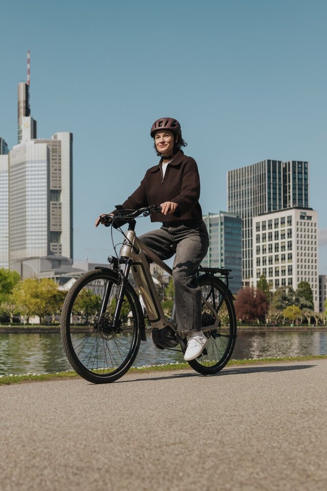 Eine Frau genießt eine Fahrt mit ihrem E-Bike auf einem Radweg am Flussufer, mit der Skyline von Frankfurt im Hintergrund.