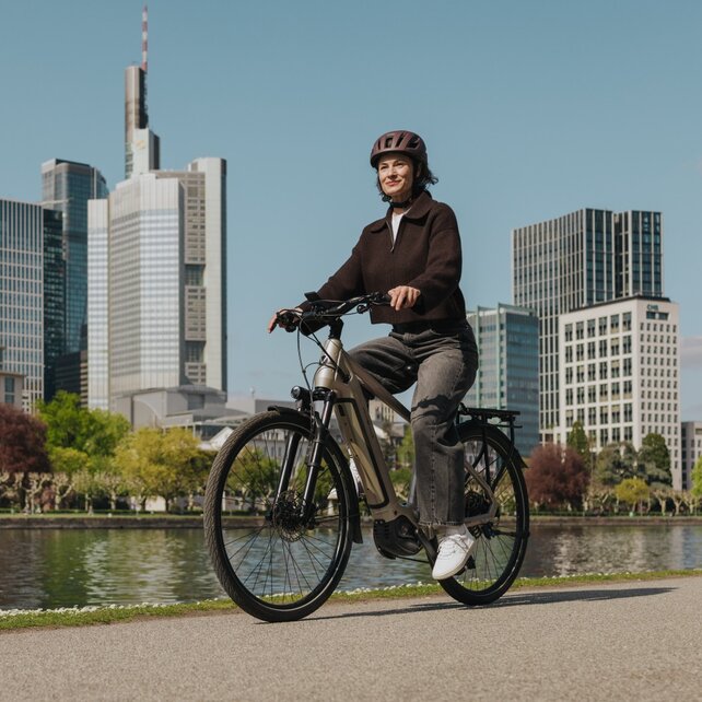 Une femme profite d'une balade en vélo électrique sur une piste cyclable au bord de la rivière, avec la silhouette de Francfort en arrière-plan.