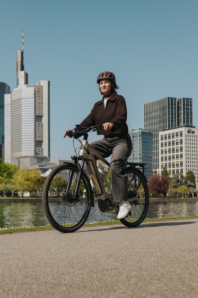 Eine Frau genießt eine Fahrt mit ihrem E-Bike auf einem Radweg am Flussufer, mit der Skyline von Frankfurt im Hintergrund.