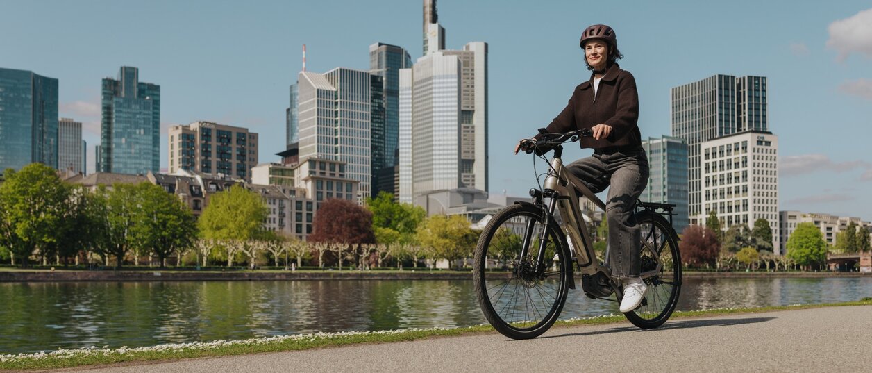 Eine Frau genießt eine Fahrt mit ihrem E-Bike auf einem Radweg am Flussufer, mit der Skyline von Frankfurt im Hintergrund.