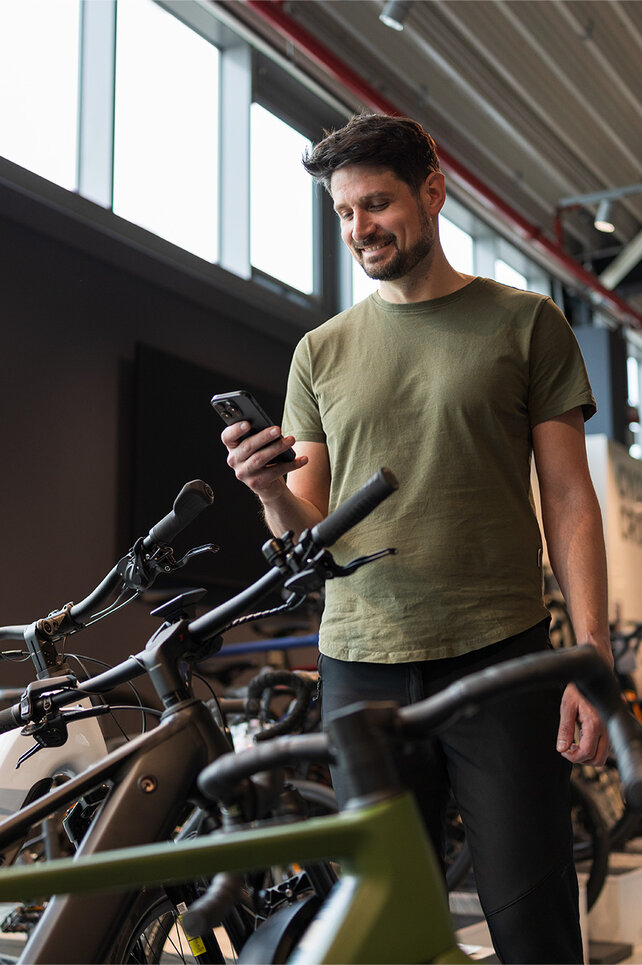 A man stands in a bike shop, smiling at his smartphone, surrounded by a variety of bikes on display.