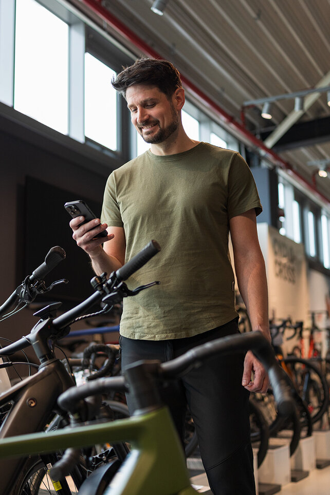 En man står i en cykelbutik, ler mot sin smartphone och är omgiven av många utställda cyklar.