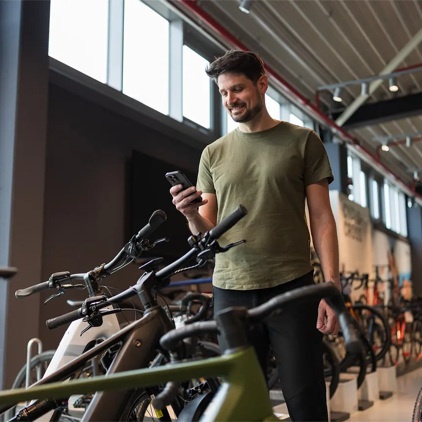 Un homme se tient dans un magasin de vélos, regarde son smartphone en souriant et se trouve entouré de nombreux vélos exposés.
