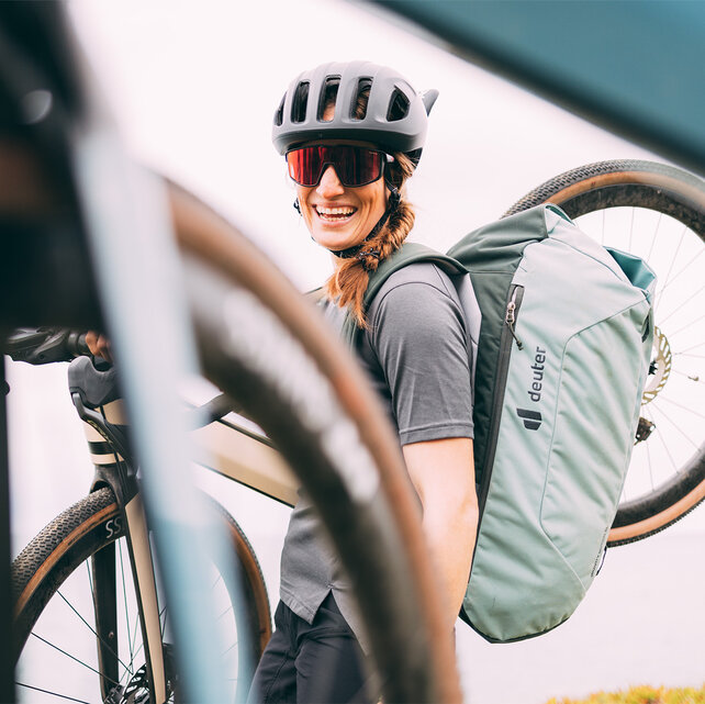 Een vrouw met een helm en een zonnebril draagt haar eGravel Bike glimlachend op haar schouder.