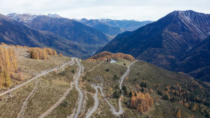 Une vue de drone montre une route en lacets qui monte vers une montagne, avec des montagnes et des arbres en arrière-plan.