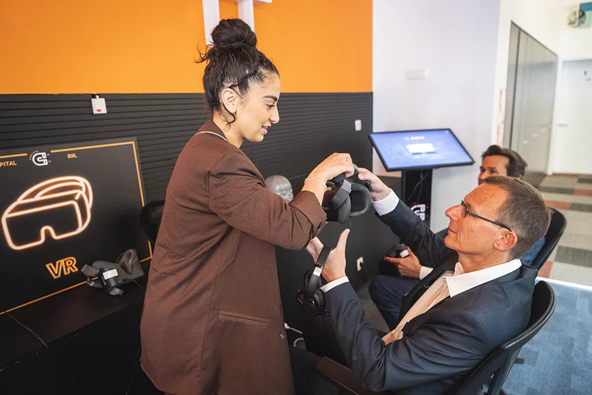 Een man in pak zit op een stoel. Voor hem hangt een bord met daarop een VR-bril en de tekst "VR". Een vrouw in pak overhandigt hem een VR-bril. Naast hem zit een andere man.