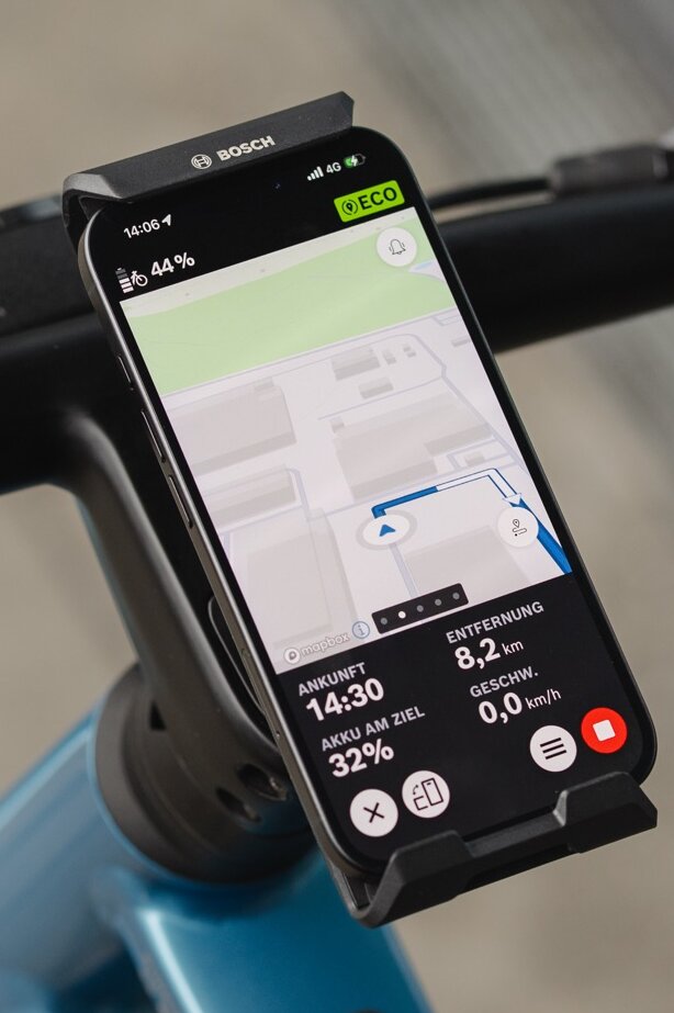 Nahaufnahme eines Smartphones, das im Bosch eBike SmartphoneGrip Vertical am Lenker befestigt ist und als eBike-Display zur Navigation genutzt wird. Die eBike Flow App zeigt eine farbige Straßenkarte mit einer blauen Routenlinie, einen bevorstehenden Abbiegehinweis nach links sowie die verbleibende Distanz und Ankunftszeit am unteren Bildschirmrand.