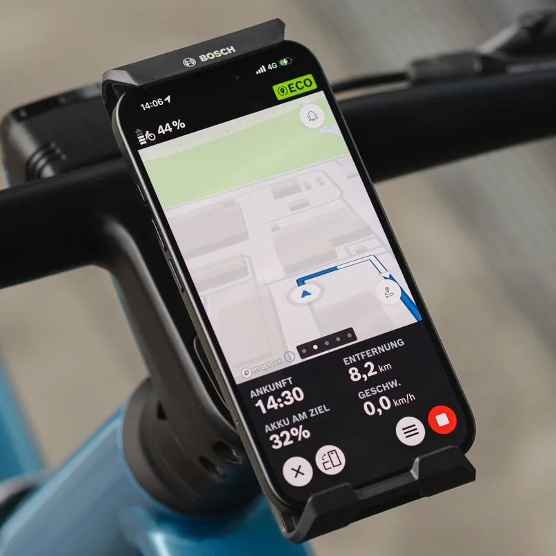 Nahaufnahme eines Smartphones, das im Bosch eBike SmartphoneGrip Vertical am Lenker befestigt ist und als eBike-Display zur Navigation genutzt wird. Die eBike Flow App zeigt eine farbige Straßenkarte mit einer blauen Routenlinie, einen bevorstehenden Abbiegehinweis nach links sowie die verbleibende Distanz und Ankunftszeit am unteren Bildschirmrand.