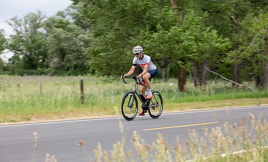 An eBike rider in short cycling gear rides along a country road bordered by trees and a pasture.