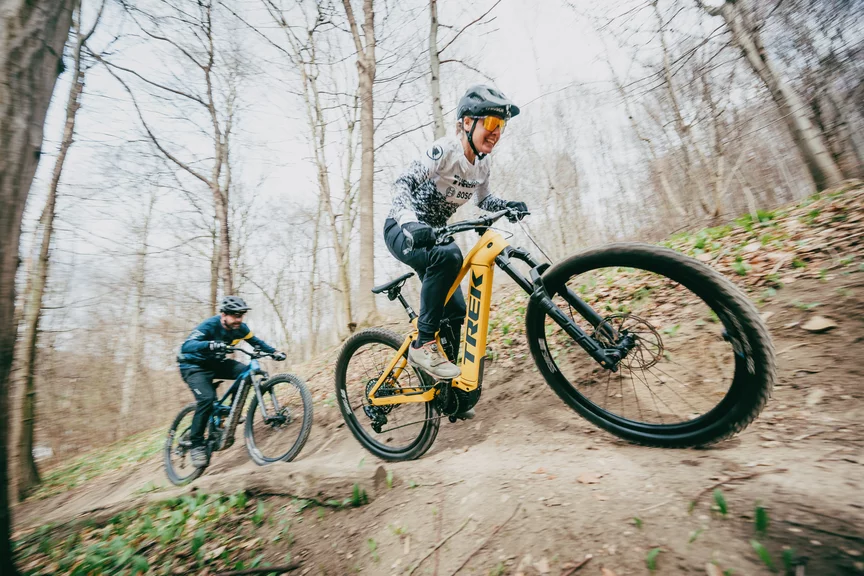 Zwei Personen fahren dicht hintereinander auf eMountainbikes einen erdigen Pfad entlang; sie lächeln freudig. Im Hintergrund sieht man kalte Baumstämme und herabgefallene braune Blätter.