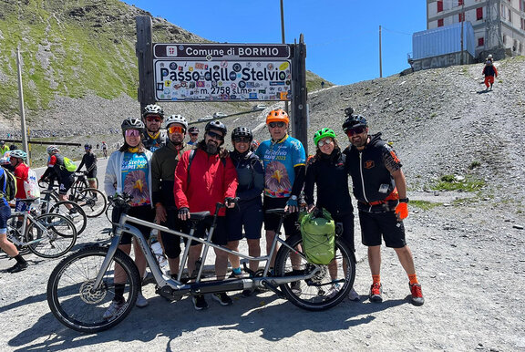 Un gruppo di ciclisti eBike posa davanti a un cartello con la scritta "Passo dello Stelvio", con un tandem davanti a loro.