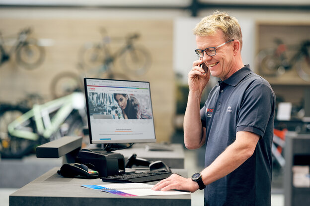 Experto en [eBike] está de pie en su escritorio en la tienda de bicicletas. En el ordenador tiene abierta la página web de Bosch mientras habla por teléfono con el Kundenservice de Bosch.