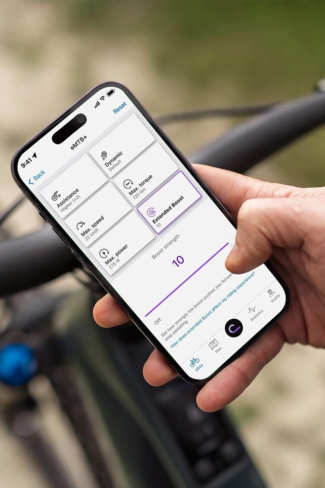 Zbliżenie na rękę trzymającą smartfon nad kierownicą e-roweru górskiego. Na wyświetlaczu jest otwarta aplikacja eBike Flow, która pozwala indywidualnie dostosować właściwości jazdy roweru elektrycznego, takie jak siła Extended Boost.
