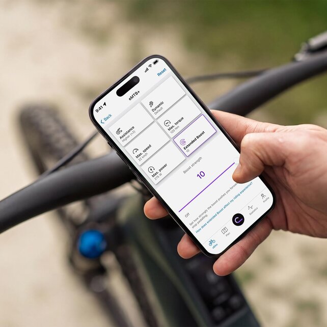 Nærbillede af en hånd, der holder en smartphone over styret på en eMountain-cykel. eBike Flow-app er åbnet på display, så du kan tilpasse elcyklens køreegenskaber individuelt – for eksempel styrken af Extended Boost.