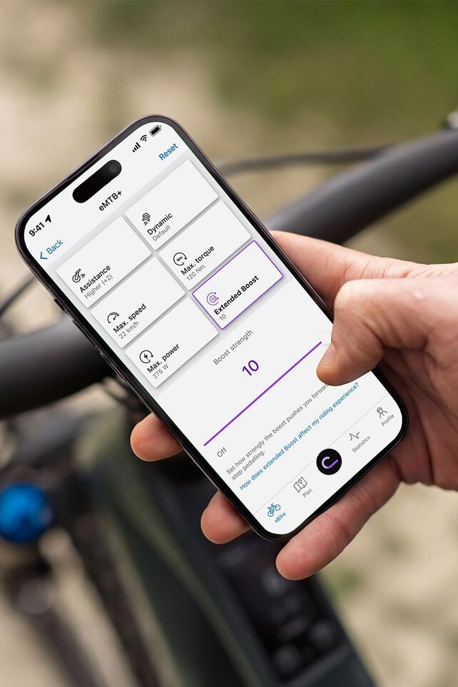 Primo piano di una mano che tiene uno smartphone sopra il manubrio di una eMountain bike. L'app eBike Flow è aperto sul display, con cui puoi regolare in modo personalizzato le caratteristiche di guida della eBike, come la resistenza della "Extended Boost".