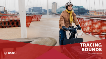 Sofie Birc rides her cargo bike across a bridge at the harbor, with cargo ships and buildings in the background. Caption: 'Tracing Sounds bosch-ebike.com'.
