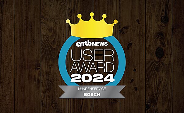 Icon mit dem Text „emtb NEWS USER AWARD 2024“: Eine goldene Krone über der Schrift, umrandet von einem blauen Kreis. Unten steht „KUNDENSERVICE BOSCH“. Der Hintergrund ist aus dunklem Holz.