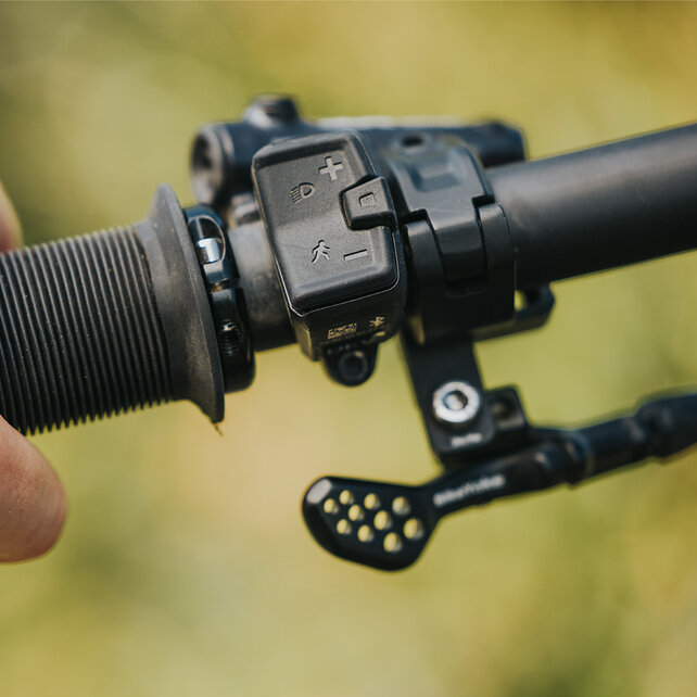 A close-up of a hand gripping a bicycle handlebar. Mounted on the handlebar is a Mini Remote from Bosch eBike Systems. The blurred background reveals an outdoor setting.