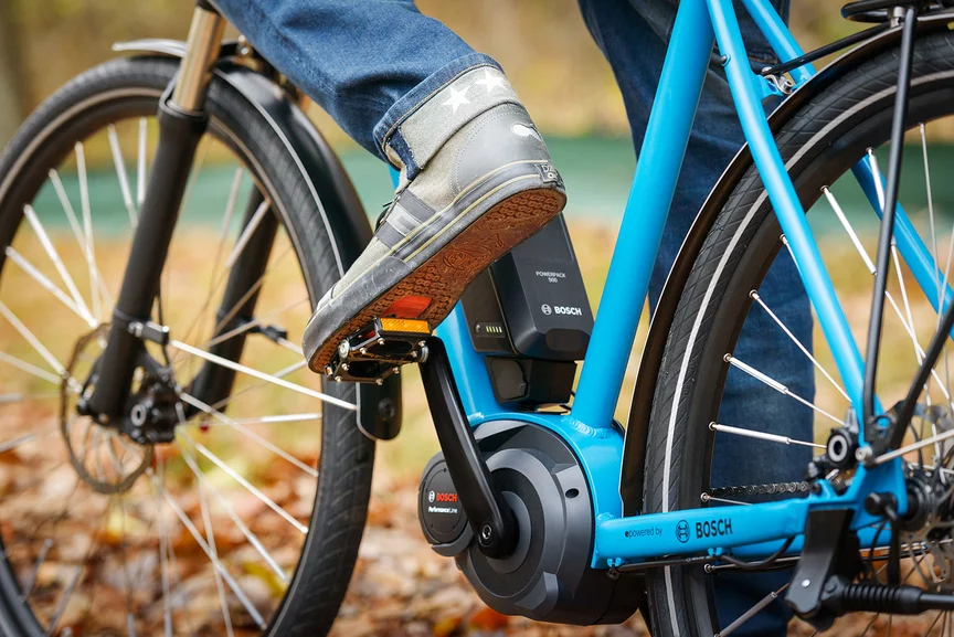 Close-up van de pedalen en aandrijfeenheid van een blauwe e-bike, waarop een persoon begint te fietsen.