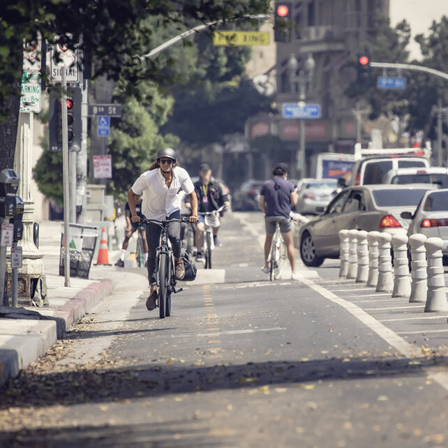 A front view of a man wearing a white shirt, black trousers, and a black helmet, riding a black eBike along a bike lane in the city. In the background, you can spot other cyclists, cars, and city buildings.
