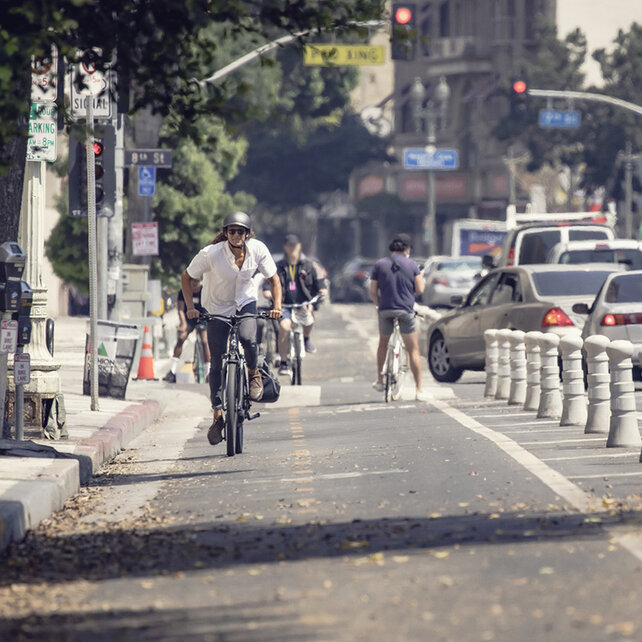A front view of a man wearing a white shirt, black trousers, and a black helmet, riding a black eBike along a bike lane in the city. In the background, you can spot other cyclists, cars, and city buildings.
