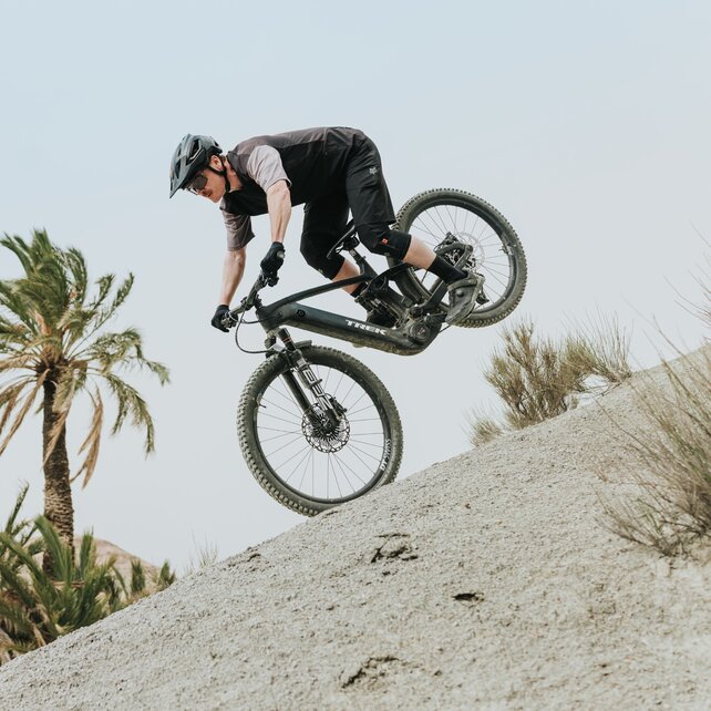 Ein Mountainbiker in voller Ausrüstung bei einem dynamischen Manöver an einem steilen Sandhang. Das Hinterrad seines eBikes ist in der Luft, während im Hintergrund Palmen und eine karge Hügellandschaft zu sehen sind.