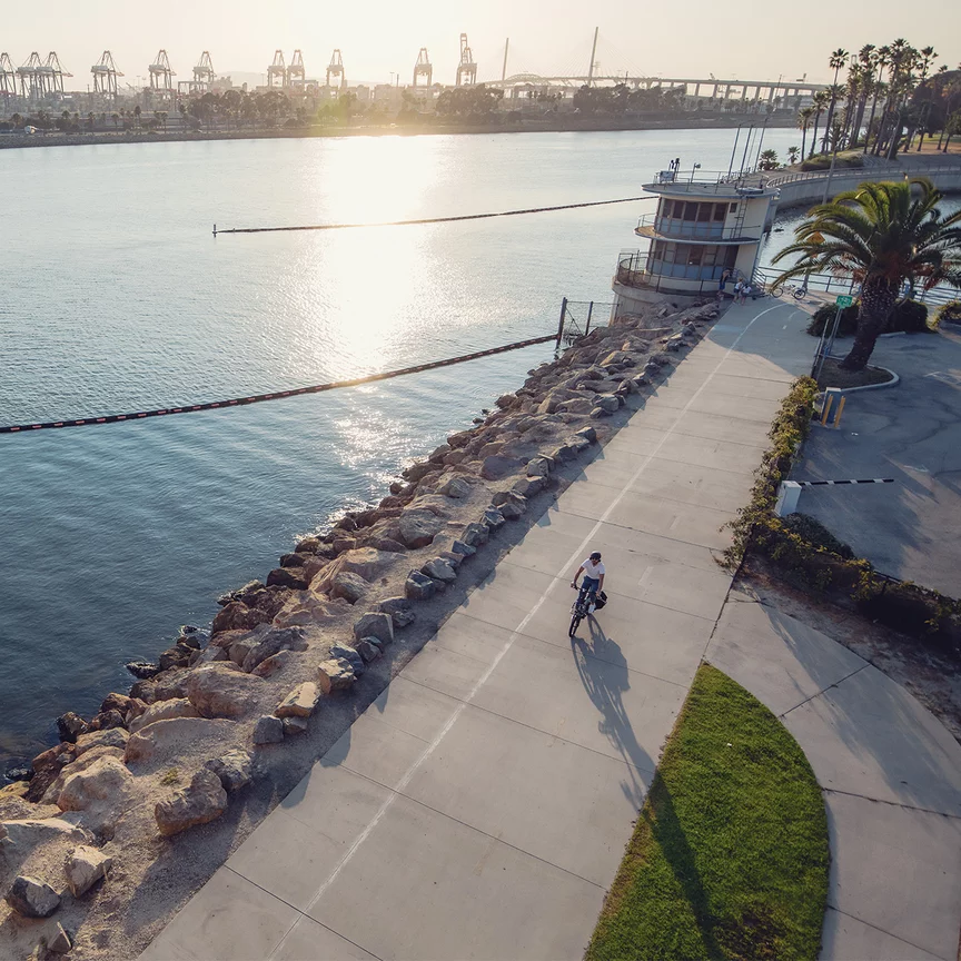 A shot from above shows a person riding an e-bike along a paved path next to the water. The path follows a rocky shoreline. In the background, harbour cranes and a bridge are visible, with the sun reflecting off the water. The scene evokes a peaceful, urban setting by the waterfront.