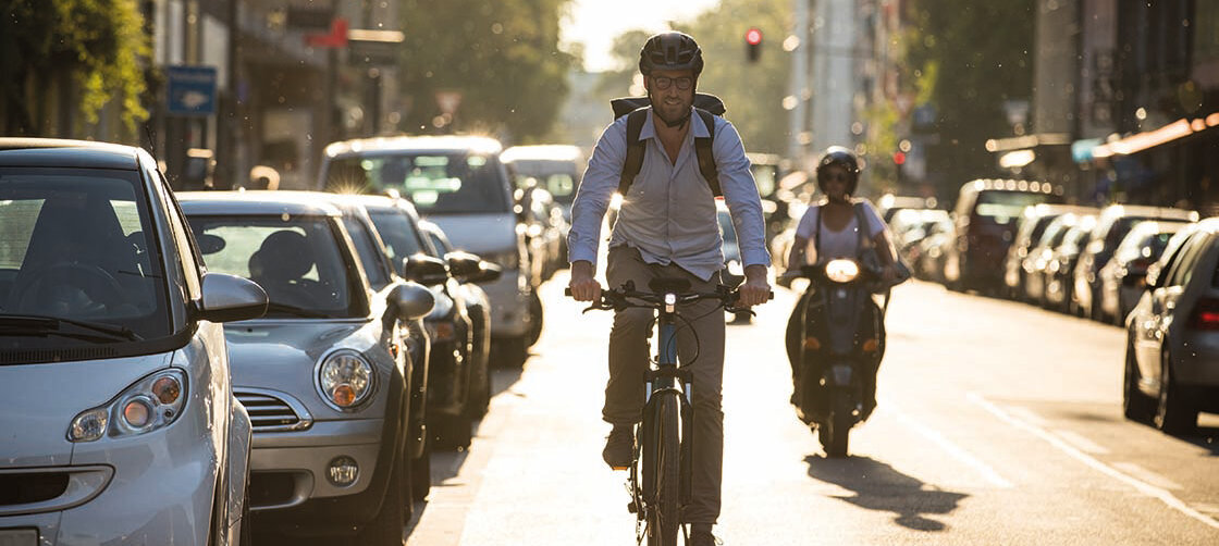 eBike rider travels along a road, with a motorcyclist behind him and parked cars to his left.