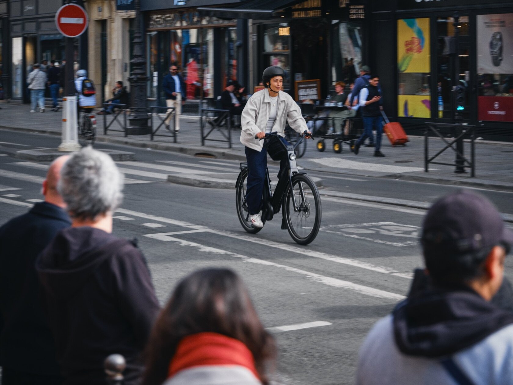 Un ciclista con casco circula en una eBike negra por una calle concurrida de la ciudad. La imagen, capturada desde la perspectiva de los transeúntes desenfocados en primer plano, muestra un momento cotidiano en el tráfico urbano entre tiendas y otras personas.
